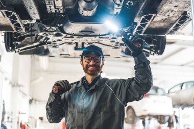 Professional car mechanic under a car on a lift using an LED lamp and repair tools. High-quality photo