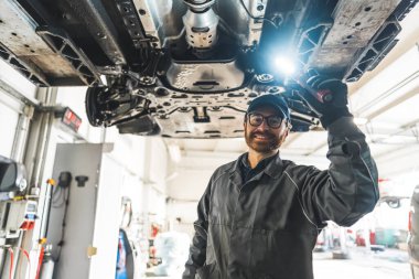 Skilled mechanic under a car on a lift using an LED lamp for chassis inspection. High-quality photo