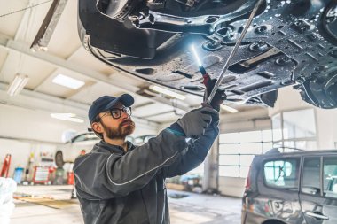 Skilled mechanic under a car on a lift using an LED lamp for chassis inspection. High-quality photo