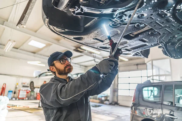 Skilled mechanic under a car on a lift using an LED lamp for chassis inspection. High-quality photo