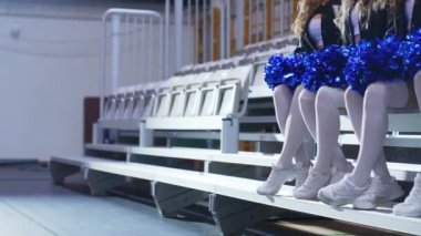 Close up side view of cheerleaders sitting in a row on a bench and holding blue-shiny pom-poms. Blurred background. High quality 4k footage