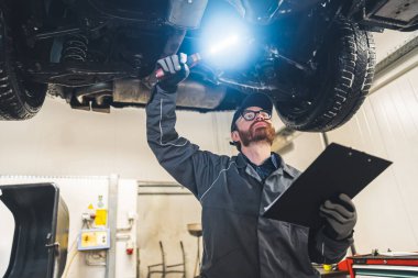 Mechanic with a clipboard inspecting the car chassis of a lifted car using a torch. High-quality photo