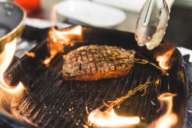 Beef striploin steak prepared on grill frying pan with herbs. Burning flames. Restaurant kitchen food preparation process. Horizontal indoor shot. High quality photo