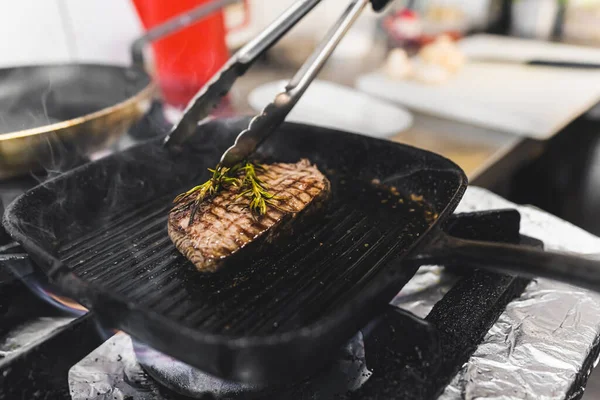 Beef striploin steak prepared on a grill frying pan with herbs. Restaurant kitchen food preparation process. Horizontal indoor shot. High quality photo