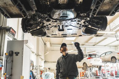 Skilled mechanic under a car on a lift using an LED lamp for chassis inspection. High-quality photo