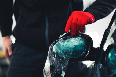 Side mirror wrapped with protective film for car paint by a professional in a red glove. Car detailing process. High quality photo
