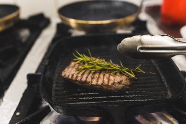 Striploin beef steak with green herbs on frying grill pan moved around with tongs. Restaurant food preparation process. Horizontal indoor shot. High quality photo