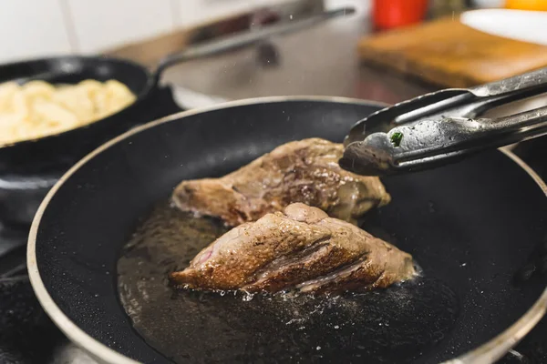Tongs flipping duck fillets frying in oil in a frying pan. Restaurant kitchen food preparation process. Horizontal indoor shot. High quality photo