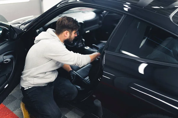 Man kneeling next to black car with open front door and cleaning leather car seats with cotton pad. Car detailing process. High quality photo
