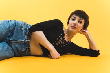 Confident short-haired woman lying on the ground and looking at camera. Low angle portrait. Studio shot over yellow background. High quality photo