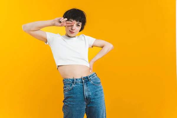 Young woman in white crop top and jeans covering her eye with two fingers. Medium studio shot over orange background. Copy space. High quality photo