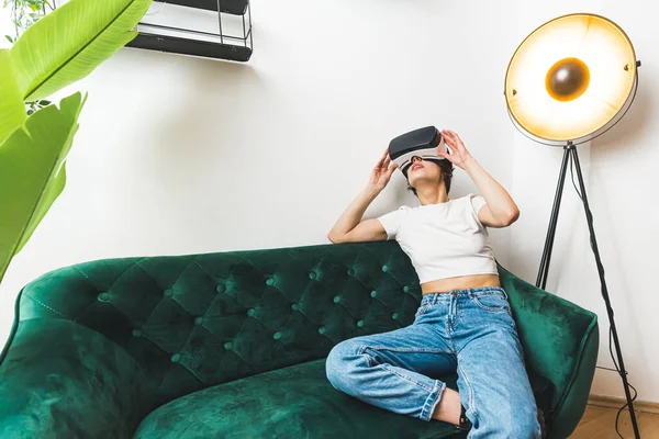 Home entertainment with modern technology. Young female gamer sitting on green sofa and enjoying video game with VR virtual reality headset. High quality photo