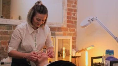 Female beautician doing a facial on her female client. The client is lying on beauty bed covered with black blanket. High quality 4k footage