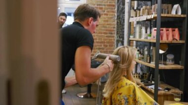 Male hairdresser uses blow dryer to style her clients blonde hair. inside the barber shop. High quality 4k footage