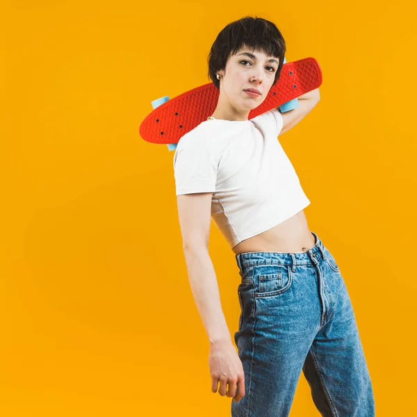 Young girl posing with small skateboard. Short-haired woman in her 20s looking at camera while holding small skateboard on her neck. Orange background. High quality photo
