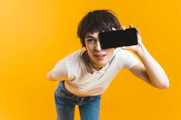 Young casually dressed woman holding phone with black screen in front of face. Copy space. Advertising. Social media. Yellow background horizontal studio shot. High quality photo