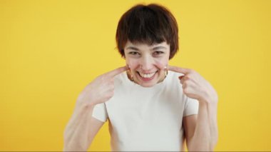 Sevimli, pozitif, genç bir kız kameraya bakıyor ve gülümseyerek gamzelerine dokunuyor. Orta boy yakın çekim. Sarı arka plan. Yüksek kalite 4k görüntü