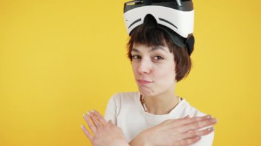 Excited smiling young adult girl looking at camera and doing silly faces with VR virtual reality headset. Medium closeup shot. Studio copy space with yellow background. High quality 4k footage