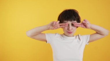 Young woman in white crop top and jeans covering her eye with two fingers. Medium studio shot over orange background. Copy space. High quality 4k footage