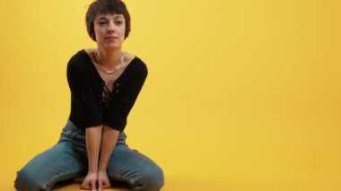 Fashionable body positive short-haired woman squatting on the ground in the studio over yellow background and looking at camera. Full-length shot. High quality 4k footage