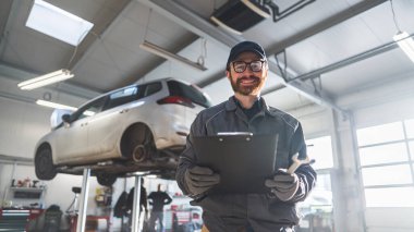Low angle shot. Mechanic holding a writing pad and smiling at the camera. Car on a lift in the background. High quality photo