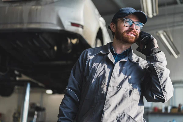 A mechanic in a repair shop talking on the phone. Medium shot. Car on the lift in the background. High quality photo