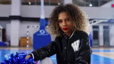 confident cheerleader with curly hair sitting on the floor with blue pom-poms in her hands. High quality 4k footage