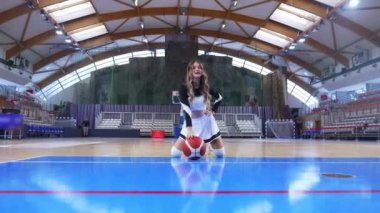 confident and cheerful cheerleader in a nice uniform kneeling and posing with a ball, indoors. High quality 4k footage