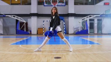 cheerful cheerleader sitting in a split on the floor with pom-poms in her hands in the arena. High quality 4k footage