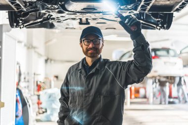 Medium shot of a mechanic posing for a picture standing under a car being held on a lift. High quality photo