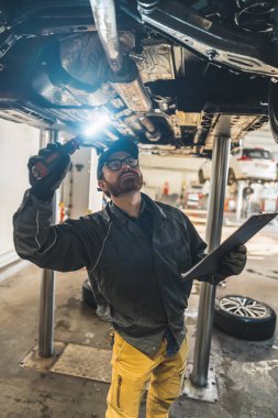 Car workshop worker checking cars chessis with a flashlight in one hand and a writing pad in another. Vertical photo. High quality photo