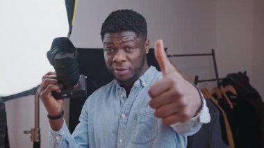 Afro-American man with a DSLR camera making thumb-up gestures and shooting a vlog in the room. High-quality 4k footage