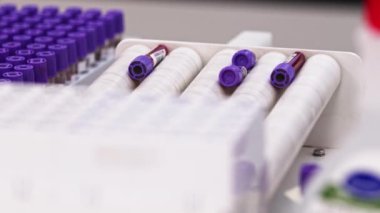 Blood samples with purple plugs being agitated on white centrifuge. Other blood samples placed in test tube holder next to the centrifuge. Blurred foreground. High quality 4k footage