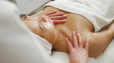 Attractive pure-skinned Asian woman in her 20s lying down on her back covered with white cotton towel during relaxing face and neck massage performed by professional massage therapist. High quality 4k