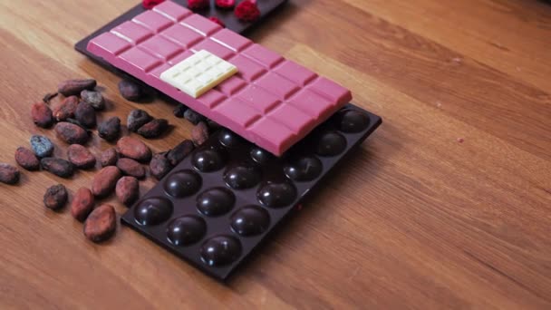 ピンクのチョコレートバーとココア豆で飾られた木製のテーブルの上に提示ミニチュアホワイトチョコレートバーと凍結乾燥ラズベリーとダークチョコレートバー プレミアムチョコレートを作る — ストック動画
