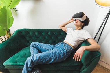 Boş zaman kavramı. Tam orta boy. Yeşil bir kanepede uzanmış, VR kulaklıkla oynayan bir kadın. Yüksek kalite fotoğraf