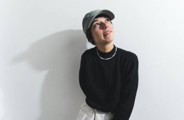 Kadın model yukarı bakıyor ve bir şapka, bir kolye ve siyah bir kazak giyiyor. Arka planda beyaz duvar var. Yüksek kalite fotoğraf