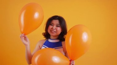 Beyaz-mavi üniformalı neşeli ponpon kız elinde turuncu balonlar, orta boy yakın çekim, stüdyo çekimi, turuncu arka plan. Yüksek kalite 4k görüntü