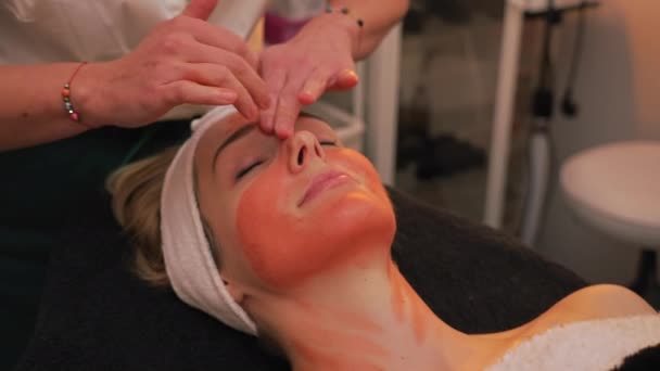 美容师用手指轻柔地将橙色黏土面罩涂在快乐的病人身上 高质量的4K镜头 — 图库视频影像