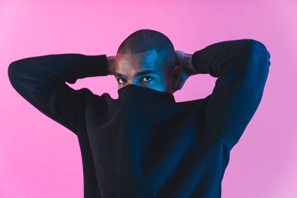 Male collage student posing for fashion photoshoot with hands behind his neck and face covered with hoodie collar. Pink background. High quality photo