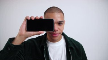 Normal giyinmiş ciddi bir erkek öğrencinin kameraya bakıp bir gözünü siyah ekran akıllı telefonla kapattığı orta boy yakın çekim. Beyaz arka plan. İzole fotokopi çekimleri. Yüksek kalite 4k görüntü