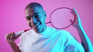 Badminton raket stüdyosu şutu çeken adam. Boynunun arkasında badminton teçhizatı tutan ve pembe arka planda kameraya bakan gülümseyen genç bir adam. Yüksek kalite 4k görüntü