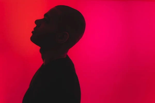 Dimmed male silhouette standing sideways to the camera and looking up against neon pink red background. High quality photo