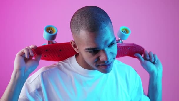 彼の首に小さなスケートボードを保持し ピンクの背景にカメラを探して白いTシャツの若い男性スケーター 高品質4K映像 — ストック動画