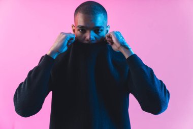 Üzgün, modaya uygun, genç bir adam yüzünü kapüşonlu bir kapüşonluyla kapatıyor ve kameraya bakıyor. Pembe arka plan. Orta boy stüdyo fotoğrafı. Yüksek kalite fotoğraf