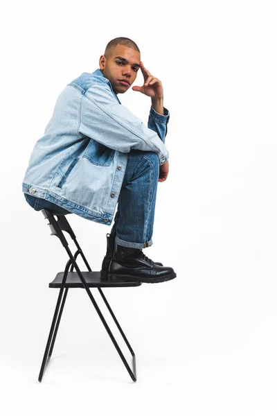 椅子の後ろに座って真剣に見てクールなデニムの服を着た若いとハンサムなアフリカ系アメリカ人の男 スタジオは垂直ショット 高品質の写真 — ストック写真