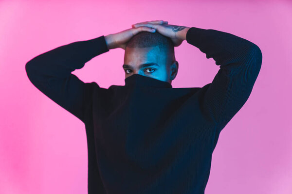 Sad Black male model with face covered with hoodie collar looking at camera over pink background. Modelling concept. High quality photo