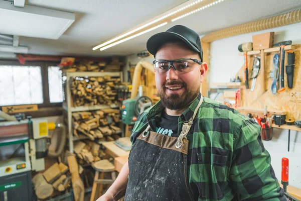 stock image Positive male carpenter in protective apron and eyewear looking at camera and smiling. Wood workshop interior. High quality photo