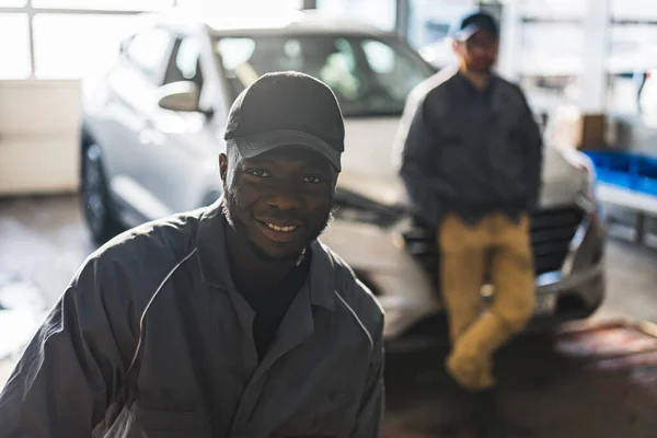 portrait of a positive African auto mechanic in uniform posing after work, he is keen on repairing cars, his co-worker in the background. High quality photo
