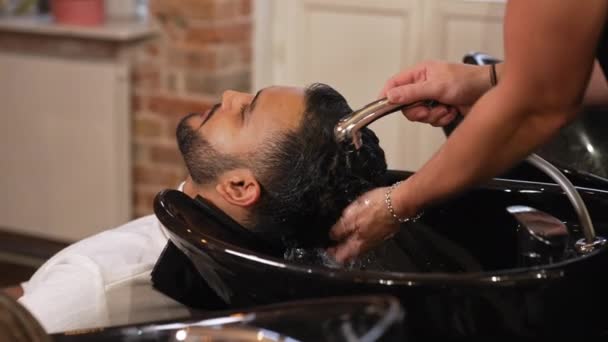 美容師はシャンプーボウルにハンドヘルドシャワーを使用してインドの男性のクライアントの髪を洗浄します 高品質4K映像 — ストック動画